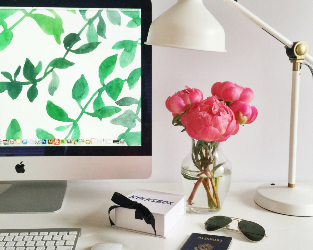 Mac on a white desk with flowers, sunglasses and passport